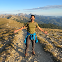 מודל גבר, לבוש ספורטיבי-הרפתקני בחולצת טי נושמת ומכנסי טיולים כהים, עומד בשביל הרים פתוח, שמש בהירה של בוקר, אווירה אנרגטית וחופשית.התמונה תכיל רק אדם אחד.