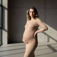 המודל ההרה, בשמלת סריג צמודה בגוון ניוד, עומדת בסטודיו מודרני עם קירות בטון חשופים, מוארת באור טבעי דרמטי, באווירה עוצמתית ואלגנטית.התמונה תכיל רק אדם אחד.